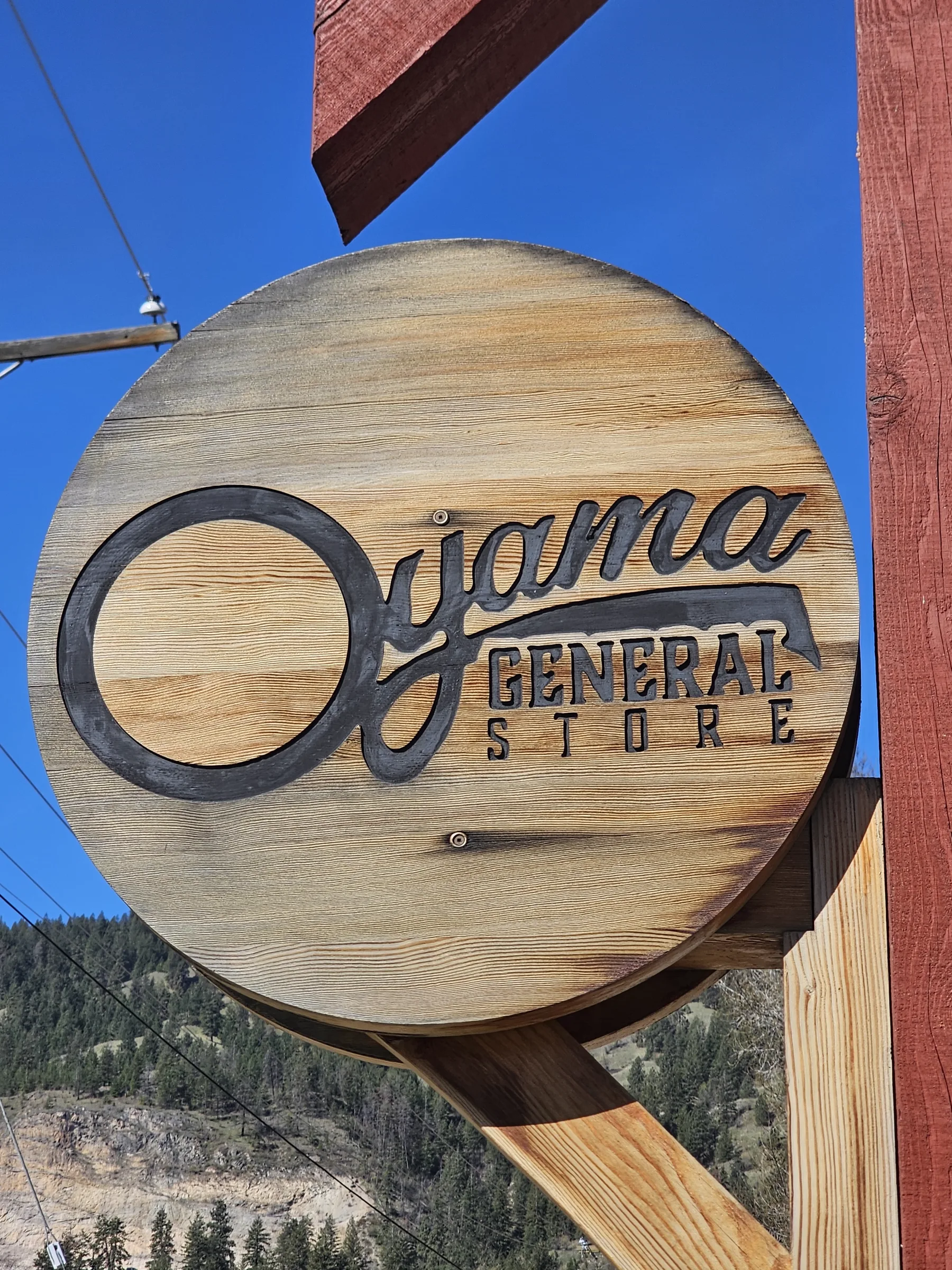 Round carved cedar Oyama General Store sign hanging against a clear blue sky