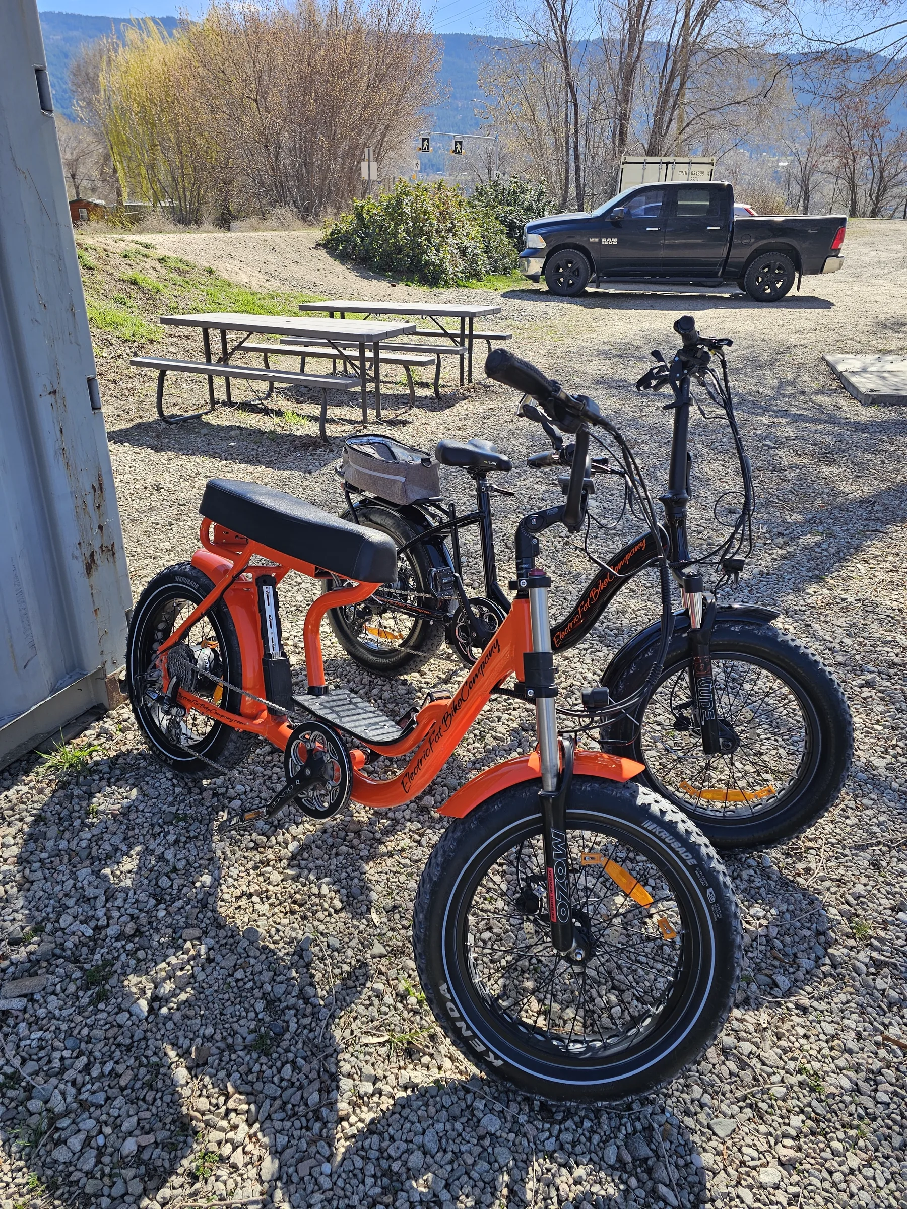 Two orange-and-black fat-tire e-bikes parked on the gravel by a picnic table and lake view